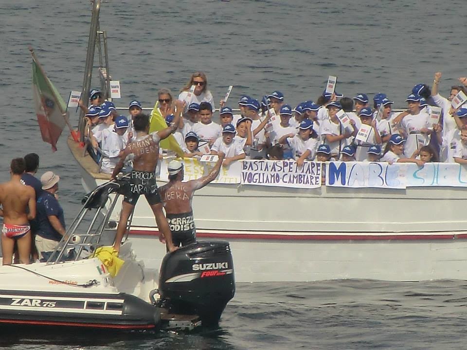 L'arrivo dei nuotatori (di spalle). Sull'imbarcazione si legge la scritta "BASTA ASPETTARE, VOGLIAMO CAMBIARE"