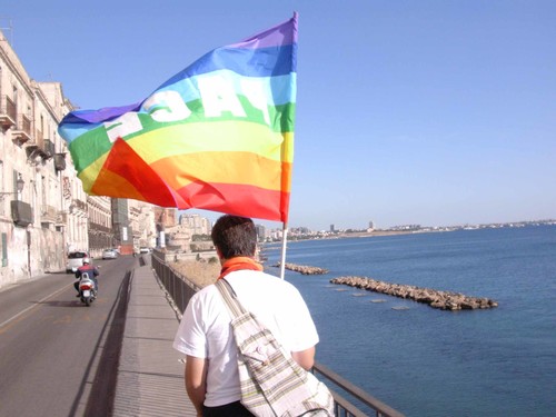 La bandiera della pace alla testa della Carovana della Pace; lungo la ringhiera dell'affaccio a mare di Taranto Vecchia camminano in fila indiana circa settanta pacifisti e di missionari. Sullo sfondo
